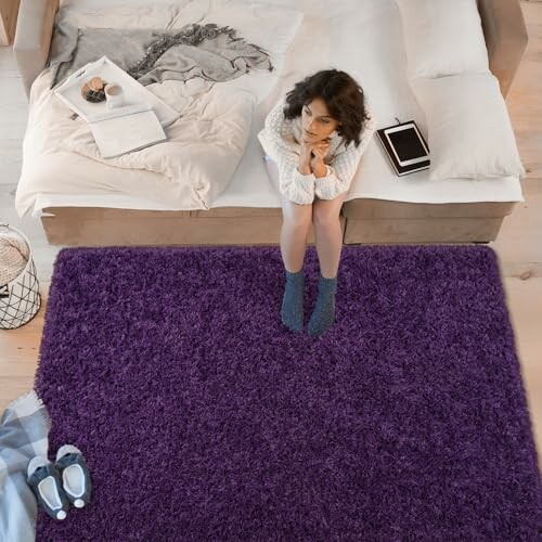 Woman sitting on bed with purple rug, book, and coffee on bed.